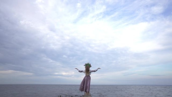站在河岸上的乌克兰年轻女子以慢动作举手向多云的天空拍摄的极宽镜头。在阴暗的夏季春日，户外穿着传统刺绣