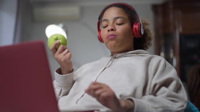 自信的非洲裔美国妇女戴着耳机在笔记本电脑上在线吃苹果短信。专注的年轻自由职业者在室内家庭办公室咬和咀