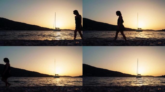 女人在金色天空下日落时与帆船一起在圆石滩上行走