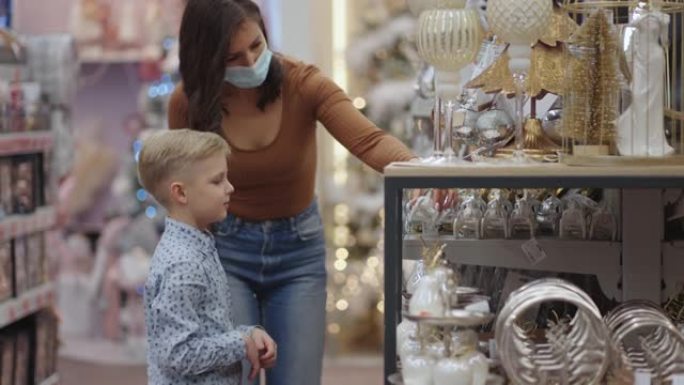 一位戴着医用口罩的母亲和她的儿子在购物中心选择圣诞节的家居装饰品。观看圣诞树玩具。冠状病毒流行病。选