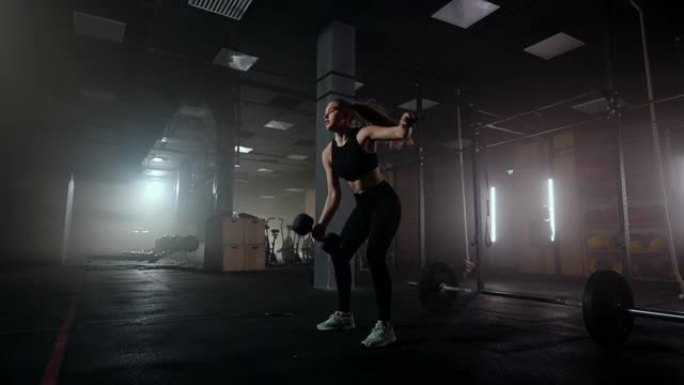 一个坚强的女人努力并克服困难在黑暗的体育馆里举起哑铃。健身女子举重哑铃在健身俱乐部训练。