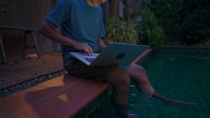 年轻人晚上用笔记本电脑在游泳池休息