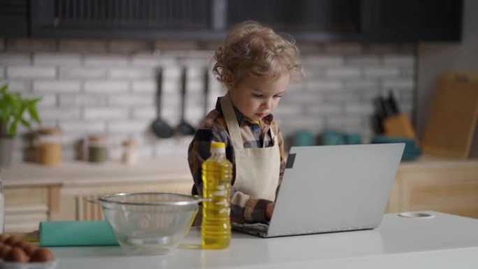 可爱的围裙蹒跚学步的孩子正在公寓的厨房里玩耍，用现代笔记本电脑准备做饭