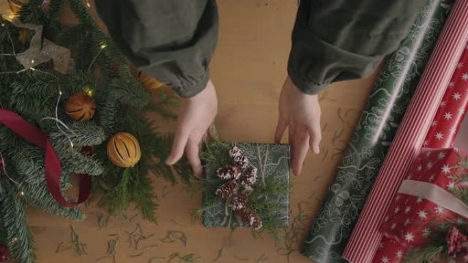 俯视图平面图。完全可见带有装饰品的桌子。双手将用手纸包裹的圣诞礼物放在木桌上并完成。包扎胶带和绑弓。