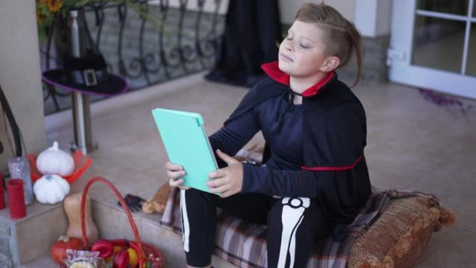 穿着吸血鬼骷髅服装的可爱的男孩坐在前院门廊上在平板电脑上在线看电影，慢动作微笑。快乐无忧无虑的高加索