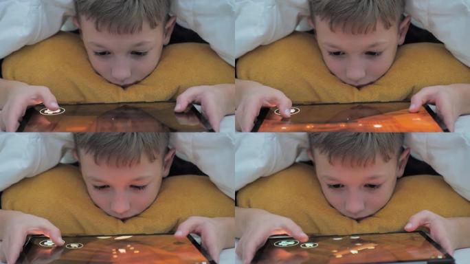 小男孩躺在毯子下在平板电脑上玩。看动画片藏起来。