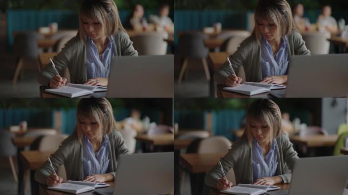 亚洲妇女在笔记本电脑上工作，坐在桌子旁写字。在咖啡店工作。女人看着笔记本电脑屏幕，在笔记本上做笔记。