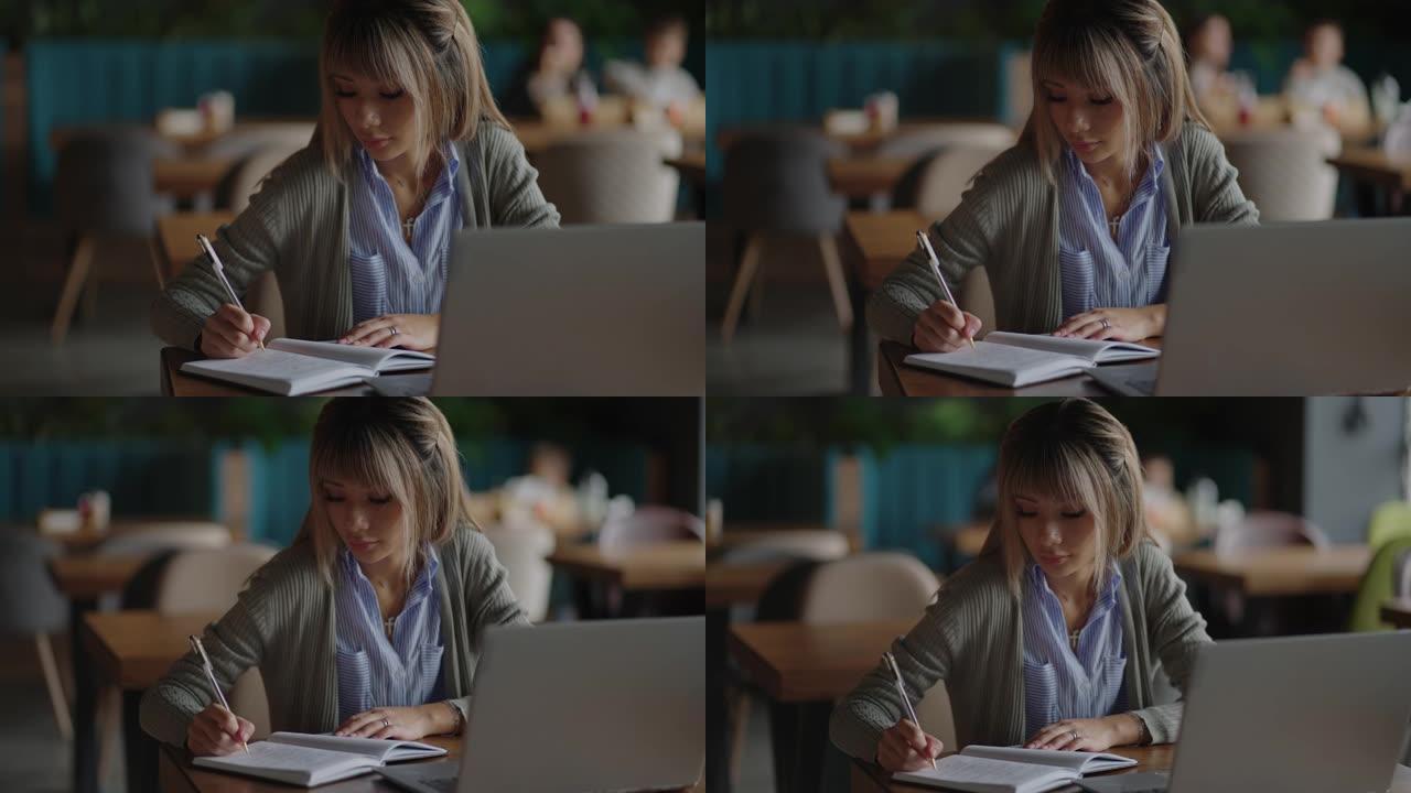 亚洲妇女在笔记本电脑上工作，坐在桌子旁写字。在咖啡店工作。女人看着笔记本电脑屏幕，在笔记本上做笔记。