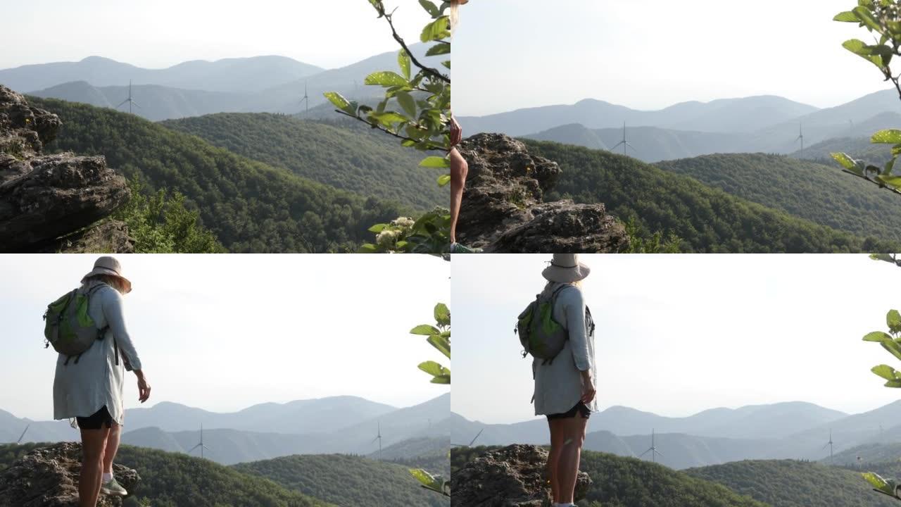 一位女徒步旅行者到达了风力涡轮机之上的岩石顶峰，绿色的山丘