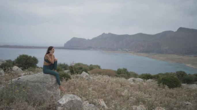 年轻女子坐在岩石上，使用手机在海滩附近的悬崖上