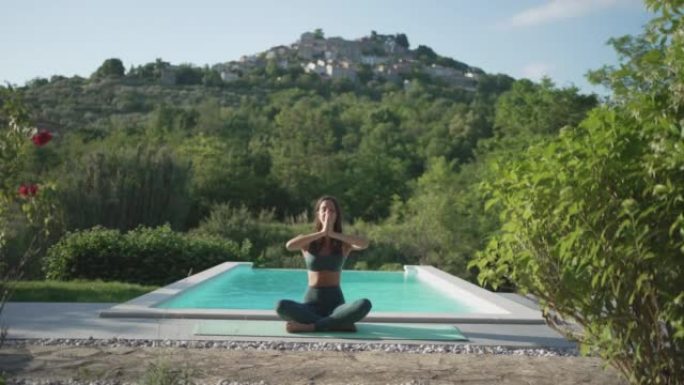 年轻女子在大自然的游泳池旁做瑜伽，俯瞰一个小村庄