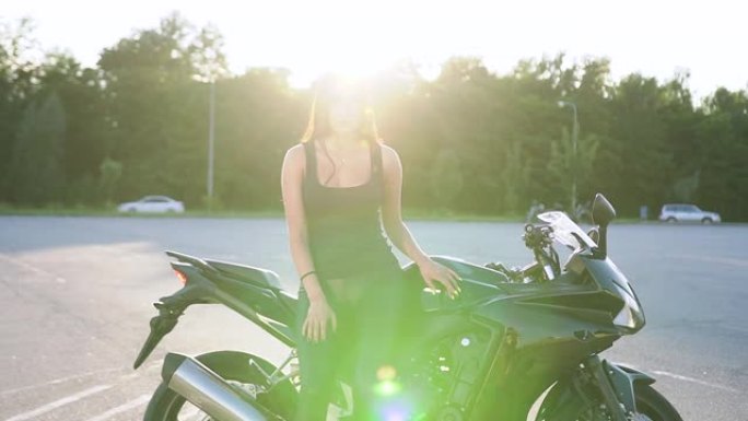 迷人时尚的年轻积极女性，穿着黑色衣服，长发，在日出下的运动场上，在她豪华的摩托车旁摆姿势