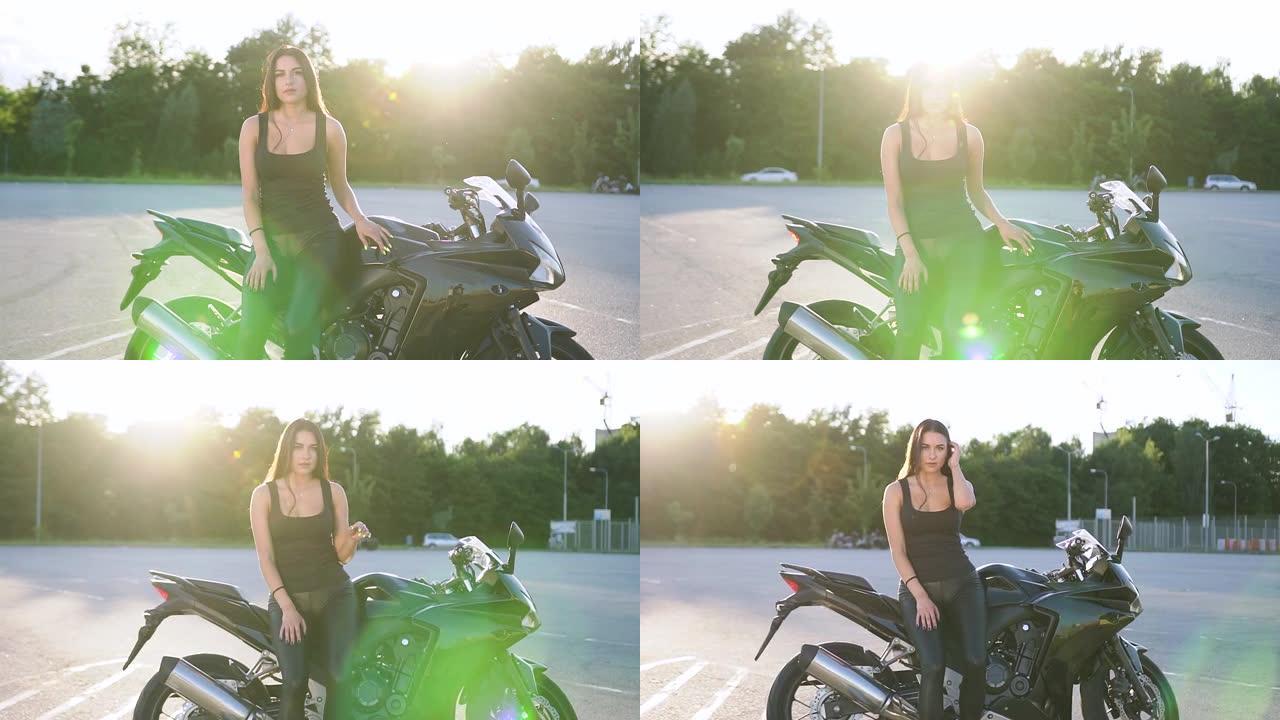 迷人时尚的年轻积极女性，穿着黑色衣服，长发，在日出下的运动场上，在她豪华的摩托车旁摆姿势