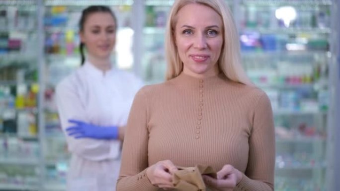 满意的女性客户的肖像在药房里与支撑长袜合影，背景模糊。前视图快乐微笑的白人妇女看着相机广告紧身衣。