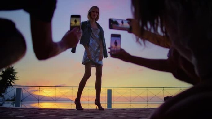 露台上浪漫的日落。美丽的女人穿着亮片连衣裙为智能手机拍照