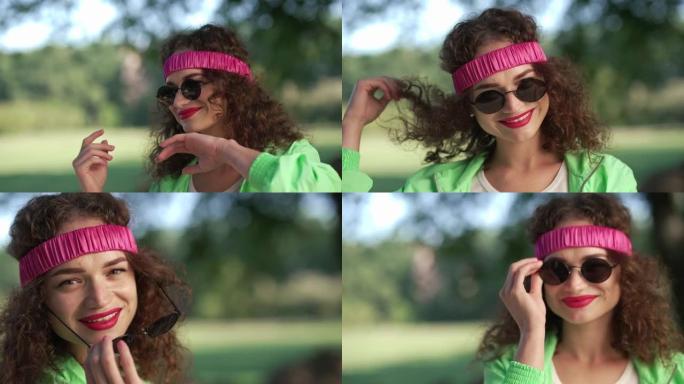 快乐自信的女人在夏日公园跳舞，放下墨镜眨眼。调情风趣的黑发卷发高加索复古女士在发带摆姿势在户外玩得开