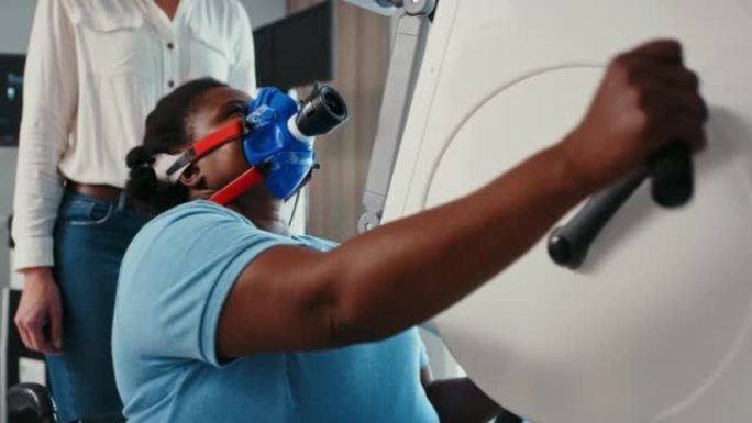 黑人妇女戴着呼吸面罩，在运动诊所或康复中心使用手踏板健身器。科学实验室用代谢分析仪测量性能和氧气水平