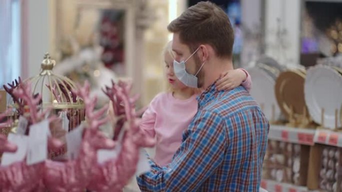 一位戴着医用口罩的父亲和他的女儿在购物中心选择圣诞节的家居装饰品。在圣诞节前夕，准备在冠状病毒大流行