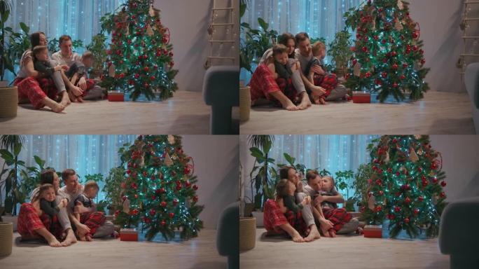 一个有两个孩子的家庭一起坐在圣诞树旁拥抱。平安夜的爱情和家庭幸福。父亲和两个儿子拥抱在客厅里看着圣诞