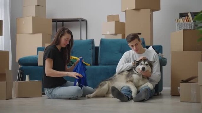 人们生活的新阶段。一个人坐在地板上，抚摸着他心爱的狗。一位朋友坐在附近，折叠衣服以搬到新家。