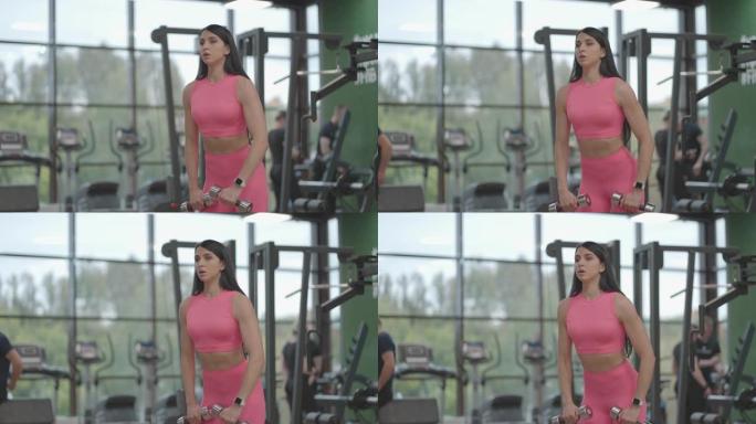 穿着粉红色西装的黑发女人在健身房训练肩膀时，交替将哑铃抬高到她面前的顶部。训练肩膀和手臂的站立练习。