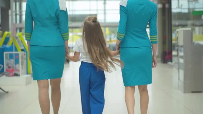 快乐的高加索女孩牵着专业空姐的手，走到机场出口。黑发孩子到达后与空乘人员漫步。陪伴概念。