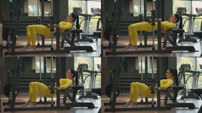 一名身穿黄色运动服的年轻女子用杠铃抬起身体，训练大腿和臀部的后部。史密斯模拟器中的重量训练。在健身房
