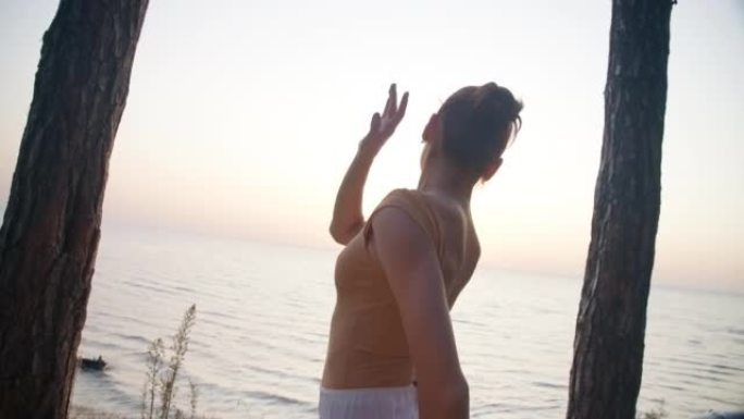 苗条优雅的芭蕾舞演员在海上夏日黎明的背景下移动双手的中拍。现场摄像机跟随才华横溢的高加索女人在日出时