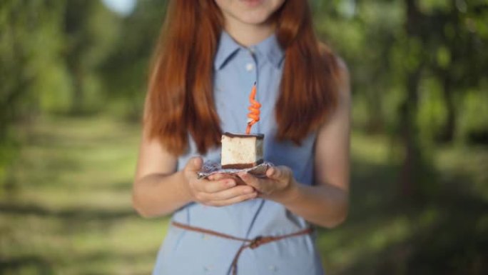 生日蛋糕放在女性青少年手中，面目全非的红发女孩站在户外吹蜡烛。快乐的高加索少年在春夏公园野餐庆祝假期