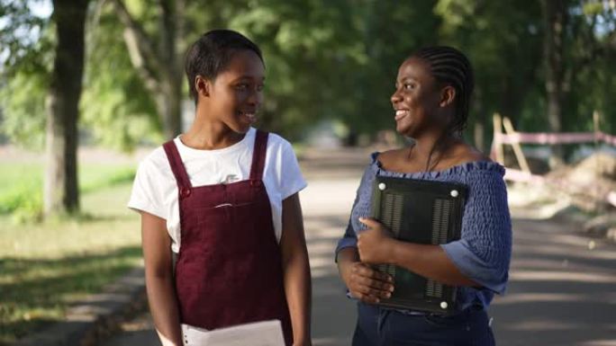 积极的非洲裔美国同伴站在阳光普照的夏日公园里大笑。中拍肖像自信聪明的年轻女性，配有笔记本电脑和户外文