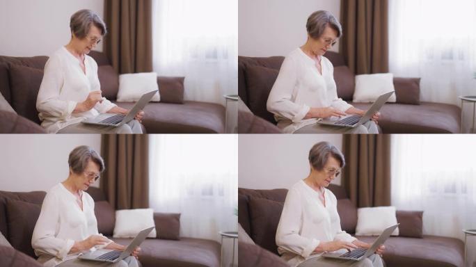 戴眼镜的高级女士使用笔记本电脑，坐在家里的沙发上，交流