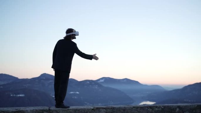 男子戴着VR虚拟现实护目镜在户外山中俯瞰日落灯光下的湖，与想象中的人握手