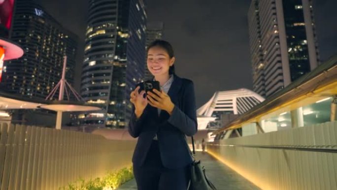 年轻的女商人开心地拿着手机走路。晚上在中心城市商业中心。