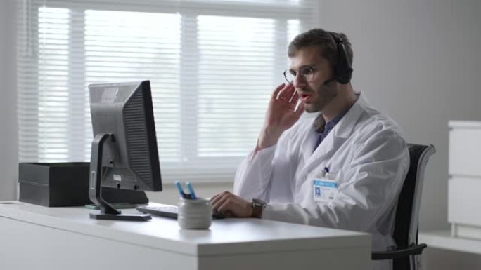 在医院工作的带耳机和计算机的男医生或护士。医疗操作员与耳机咨询患者的特写。在药品呼叫中心工作的医务人
