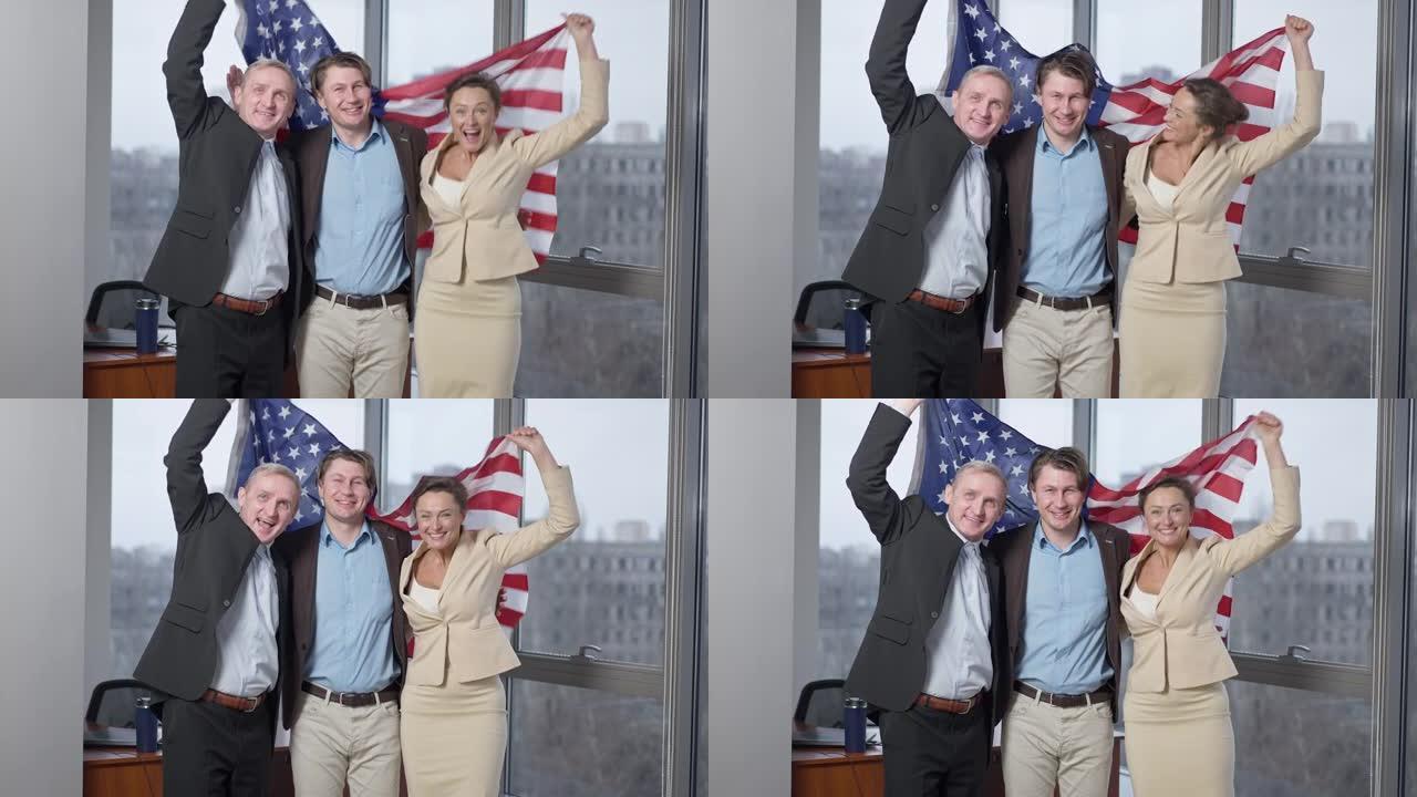 开朗的美国商业团队在办公室与国旗合影。快乐微笑的男人和女人拿着美国国家标志看着相机欢欣鼓舞的肖像。爱