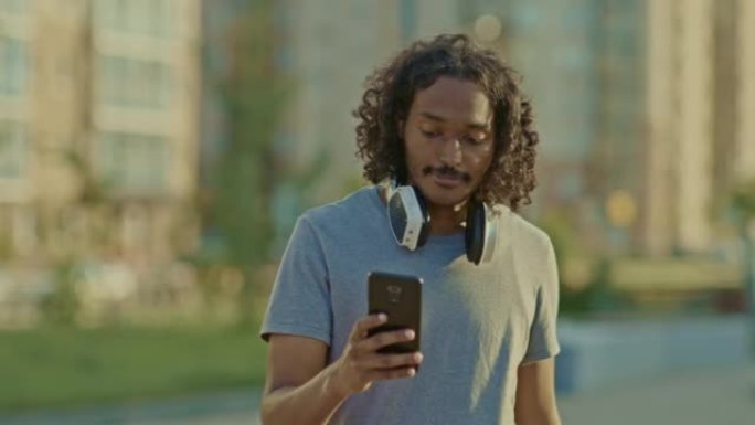 年轻人穿过城市，从口袋里掏出一部手机。皮肤黝黑的男人在手机上写字并微笑。4k，普洛雷斯
