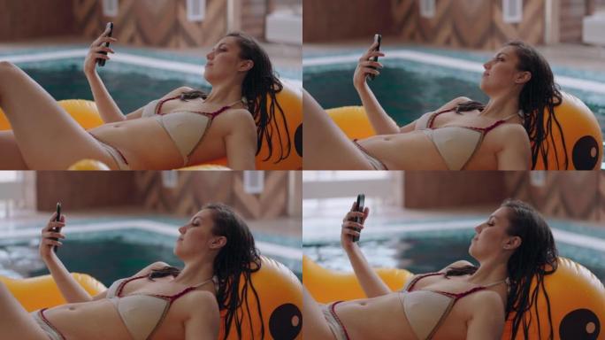穿着比基尼的成年黑发女士正在游泳池或健康中心放松，使用躺在充气圈上的智能手机