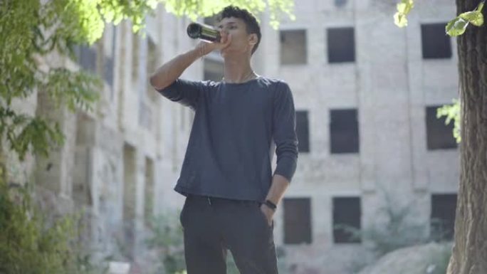 年轻的混血成瘾男子喝着瓶子里的啤酒在废墟上看着镜头。有酗酒倾向的无家可归者的肖像。虐待和贫困。