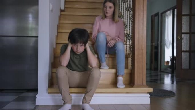 白人兄弟的宽镜头肖像用手遮住耳朵，姐姐说话时表情不满意。小男孩在室内坐在楼梯上与十几岁的女孩争吵。