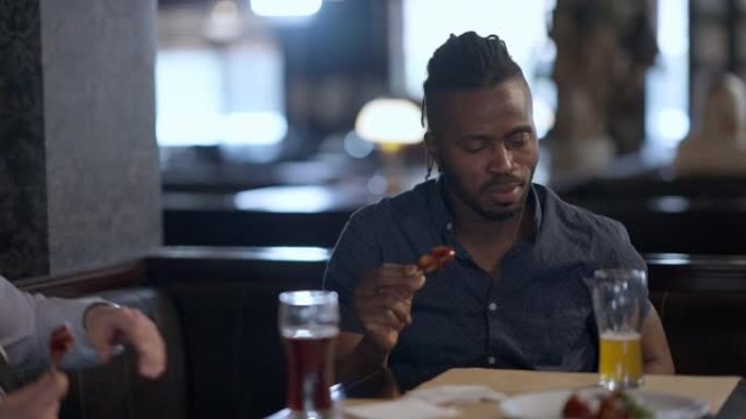 满意的非洲裔美国人的肖像与朋友在餐厅享受烤鸡喝啤酒的味道。英俊的积极客户在室内吃美味的零食。