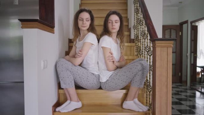 不满的双胞胎姐妹穿着睡衣背靠背坐在楼梯上。侧视肖像恼怒的高加索年轻同卵女人思考。兄弟姐妹冲突。