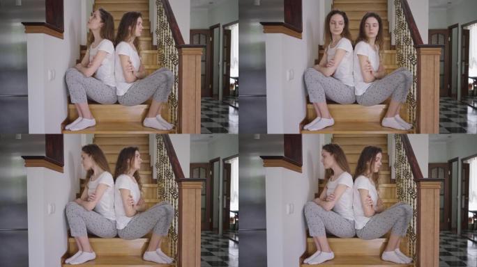 不满的双胞胎姐妹穿着睡衣背靠背坐在楼梯上。侧视肖像恼怒的高加索年轻同卵女人思考。兄弟姐妹冲突。