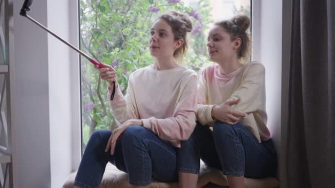 自信的双胞胎姐妹博客作者，智能手机坐在窗台上，在线聊天微笑。开朗苗条快乐的年轻白人女性在家室内聊天。