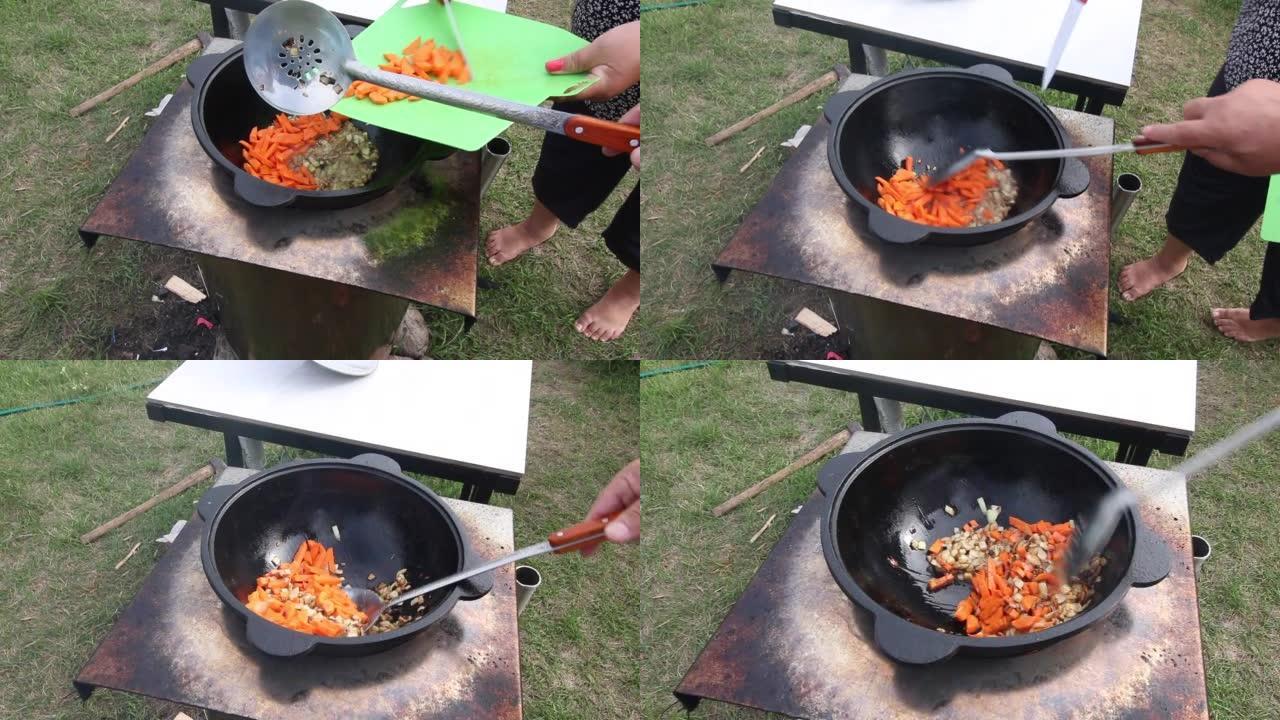 一个男人在铸铁大锅里炸洋葱。女人给它加了胡萝卜。在户外准备食物。