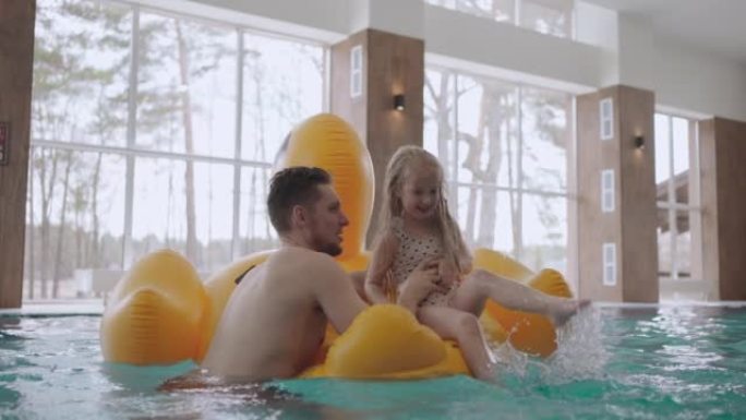 成年男子和他的小女儿在游泳池里游泳，小女孩坐在有趣的充气鸭上