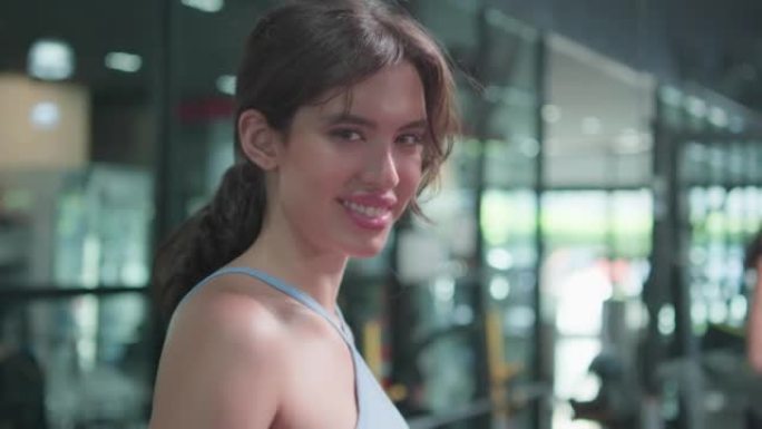 迷人的年轻女子在健身房锻炼时从瓶子里喝水快乐的笑脸。适合运动员的漂亮女孩穿着运动服，手里拿着疲惫的水