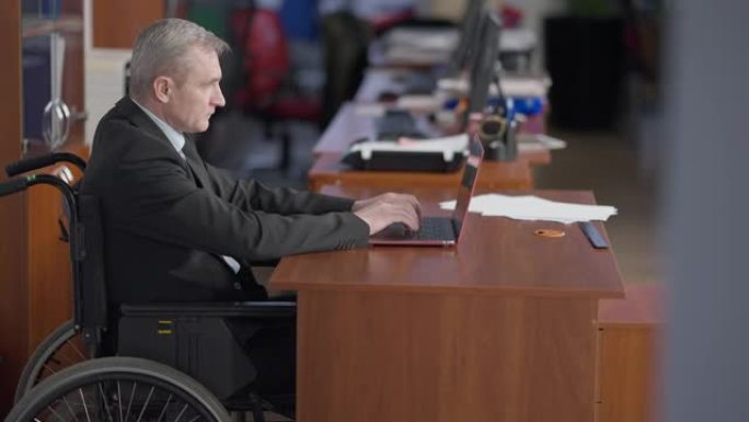 坐在办公室里用笔记本电脑键盘打字的坐在轮椅上的白人男子的侧视图。专注的男性残疾员工在室内在线工作。残