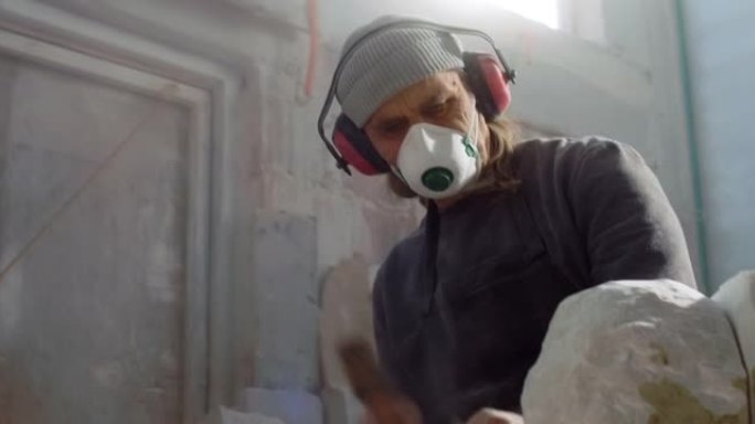 男性雕塑家在凿刻一块石头时戴着防护口罩和耳机
