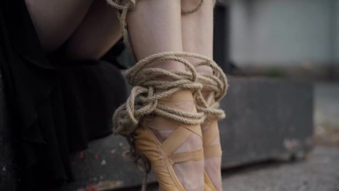 相机沿着用绳子绑住的苗条芭蕾舞女演员的腿向下移动。无法辨认的年轻白人妇女坐在户外。与职业相关的舞者概
