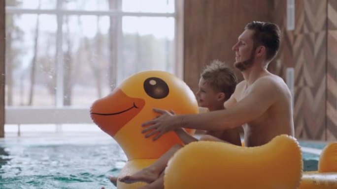 快乐的爸爸和小儿子在游泳池里骑着有趣的充气鸭，周末玩得开心，休息